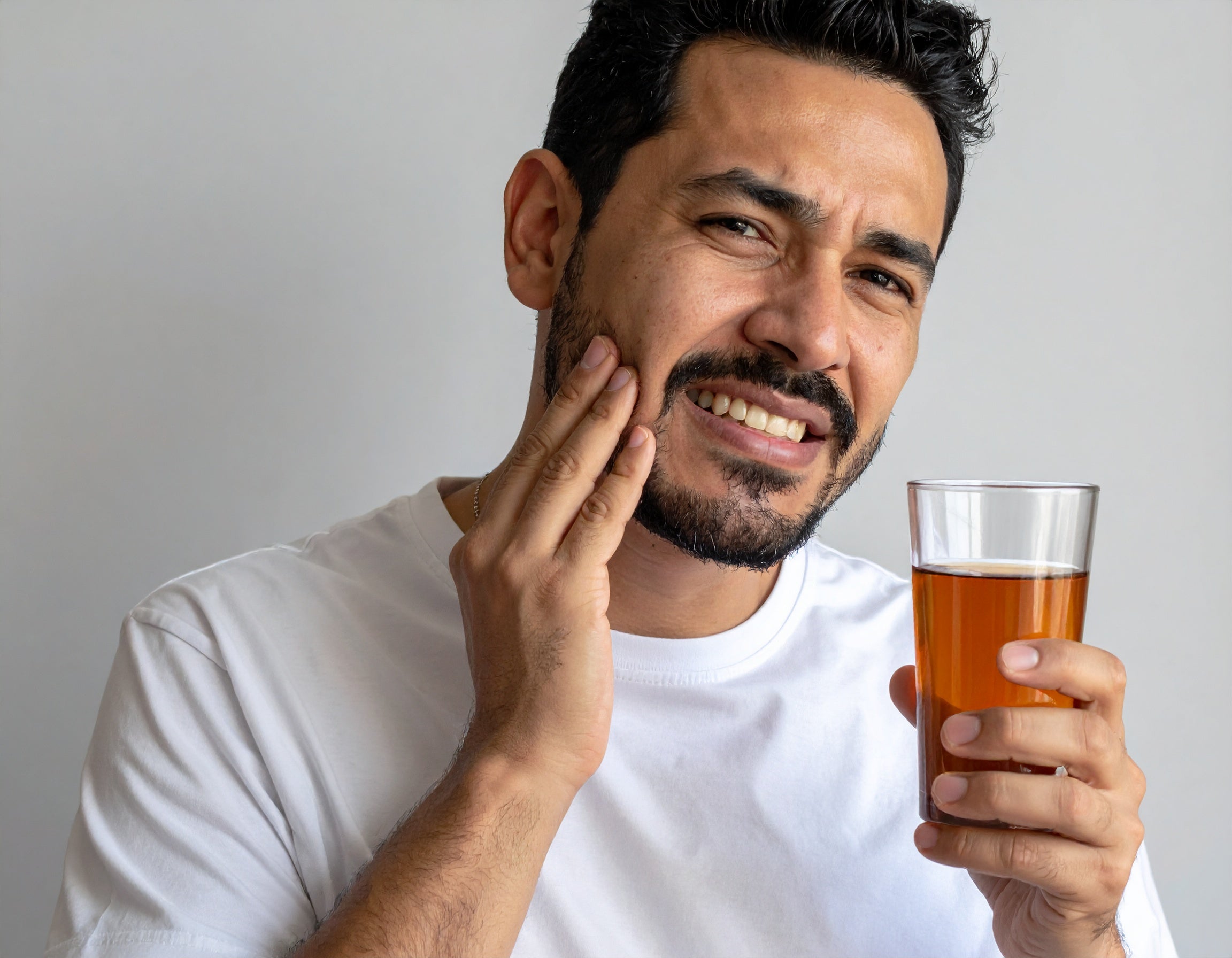 Mężczyzna chwyta policzek, ponieważ boli go ząb. W ręku trzyma zimny napój. Cierpi na nadwrażliwość zębów.